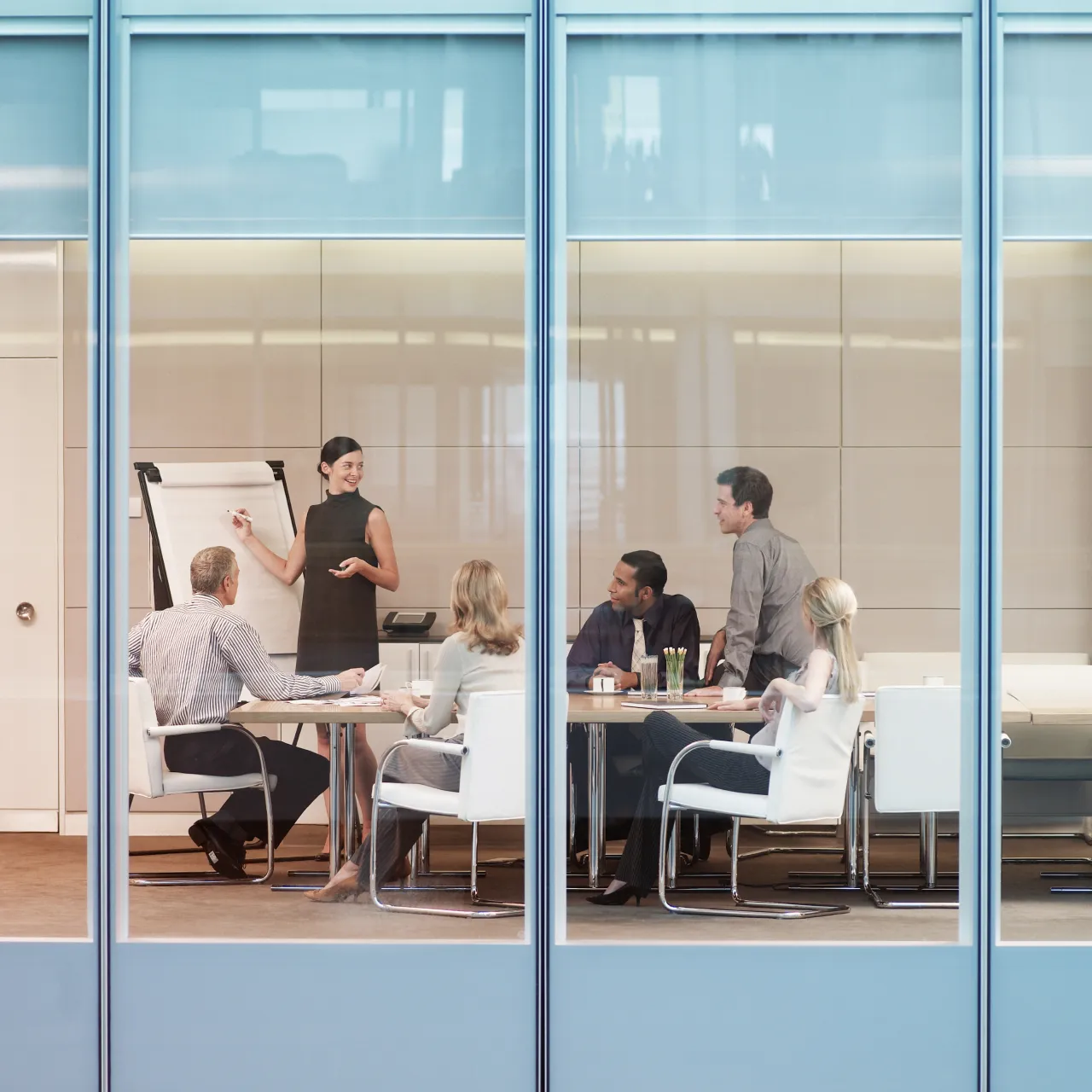 Businesswoman presenting on a flip chart to four colleagues seated around a conference table in a modern office.