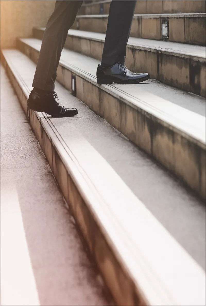 Person wearing black dress shoes and dark pants walking up concrete stairs.