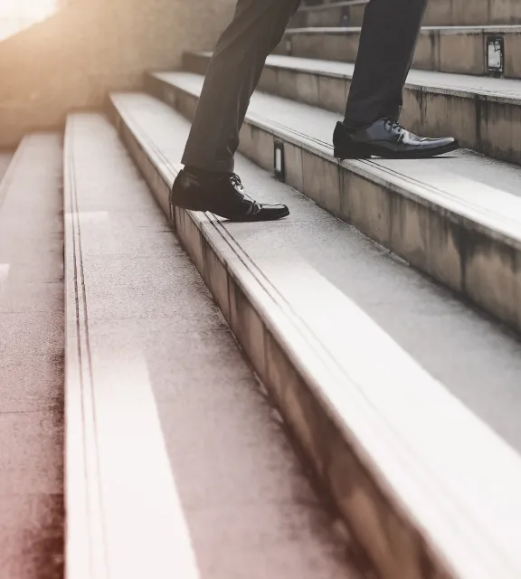 Person wearing black dress shoes and black pants walking up outdoor stone steps in sunlight.