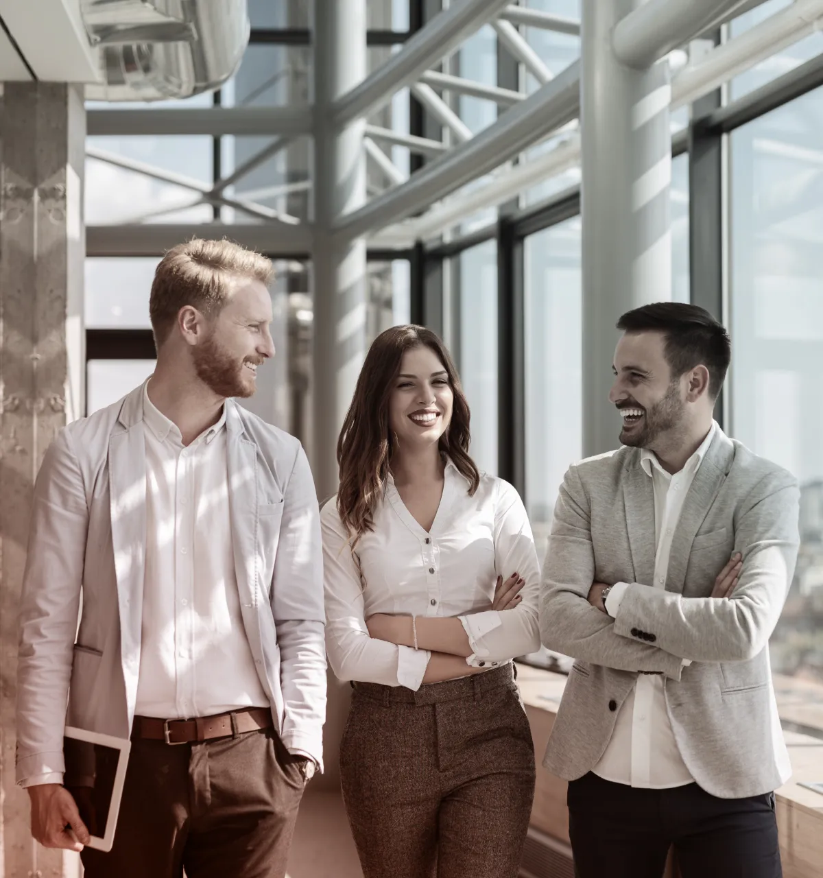 Three coworkers standing together in a bright office hallway, smiling and engaged in conversation.