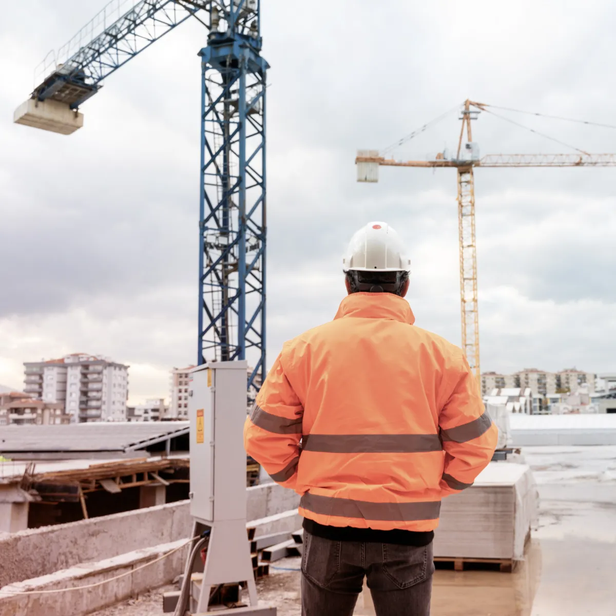Construction worker in an orange safety jacket and white hard hat overlooking a building site with cranes and apartment buildings in the background.