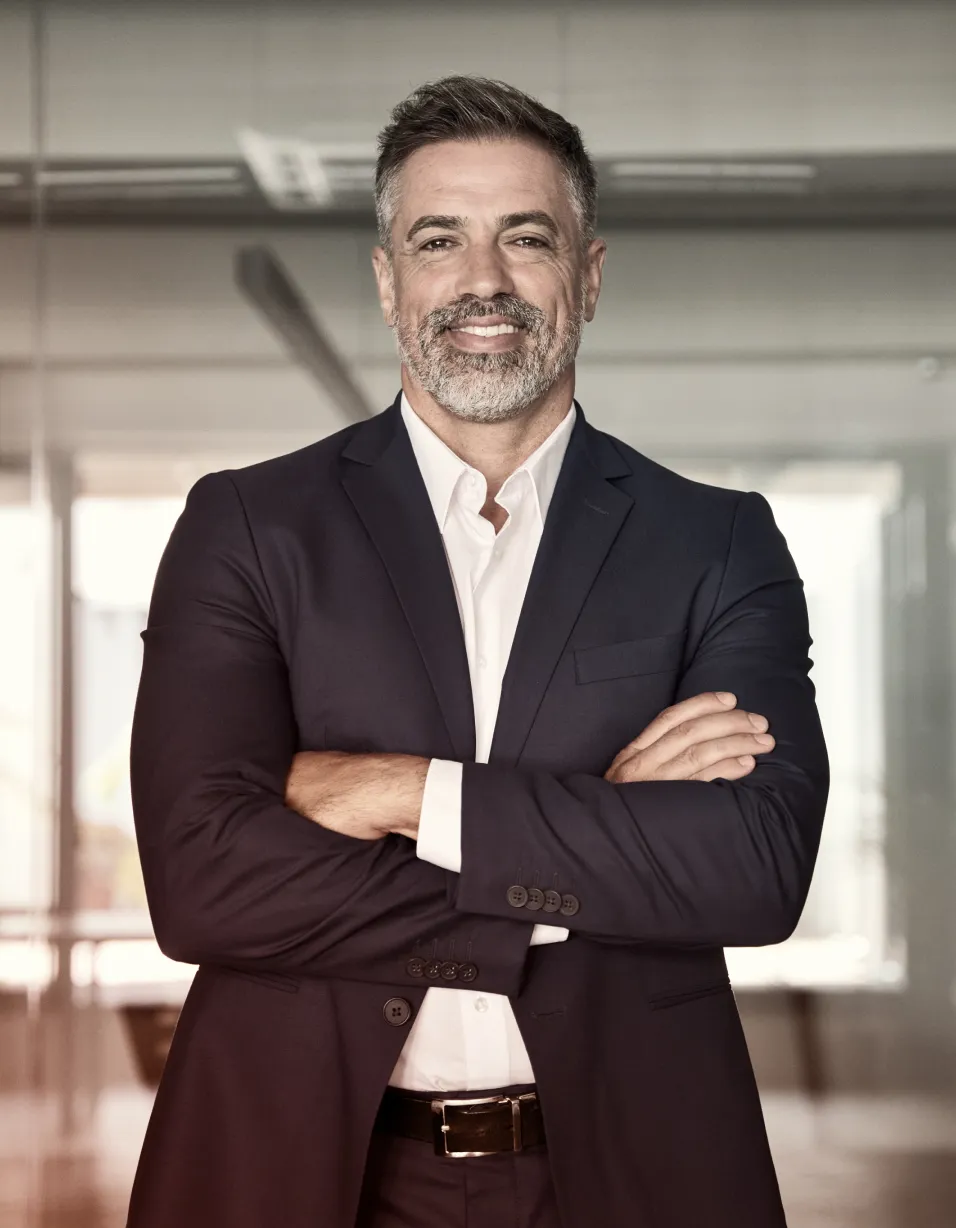 Smiling middle-aged man with gray hair and beard wearing a dark suit and white shirt, standing with arms crossed in a modern office.