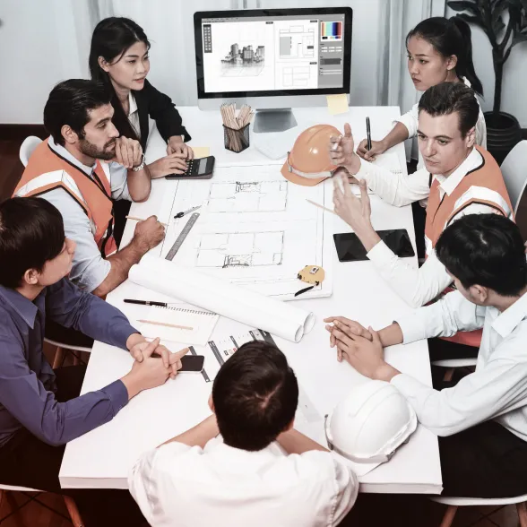 Group of architects and engineers discussing building plans around a table with blueprints and a computer monitor displaying architectural drawings.