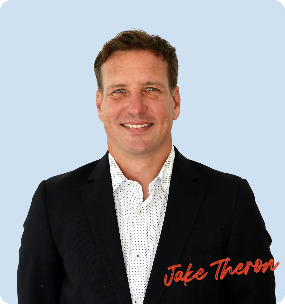Smiling man with short brown hair wearing a black suit jacket and white dotted shirt against a light blue background.