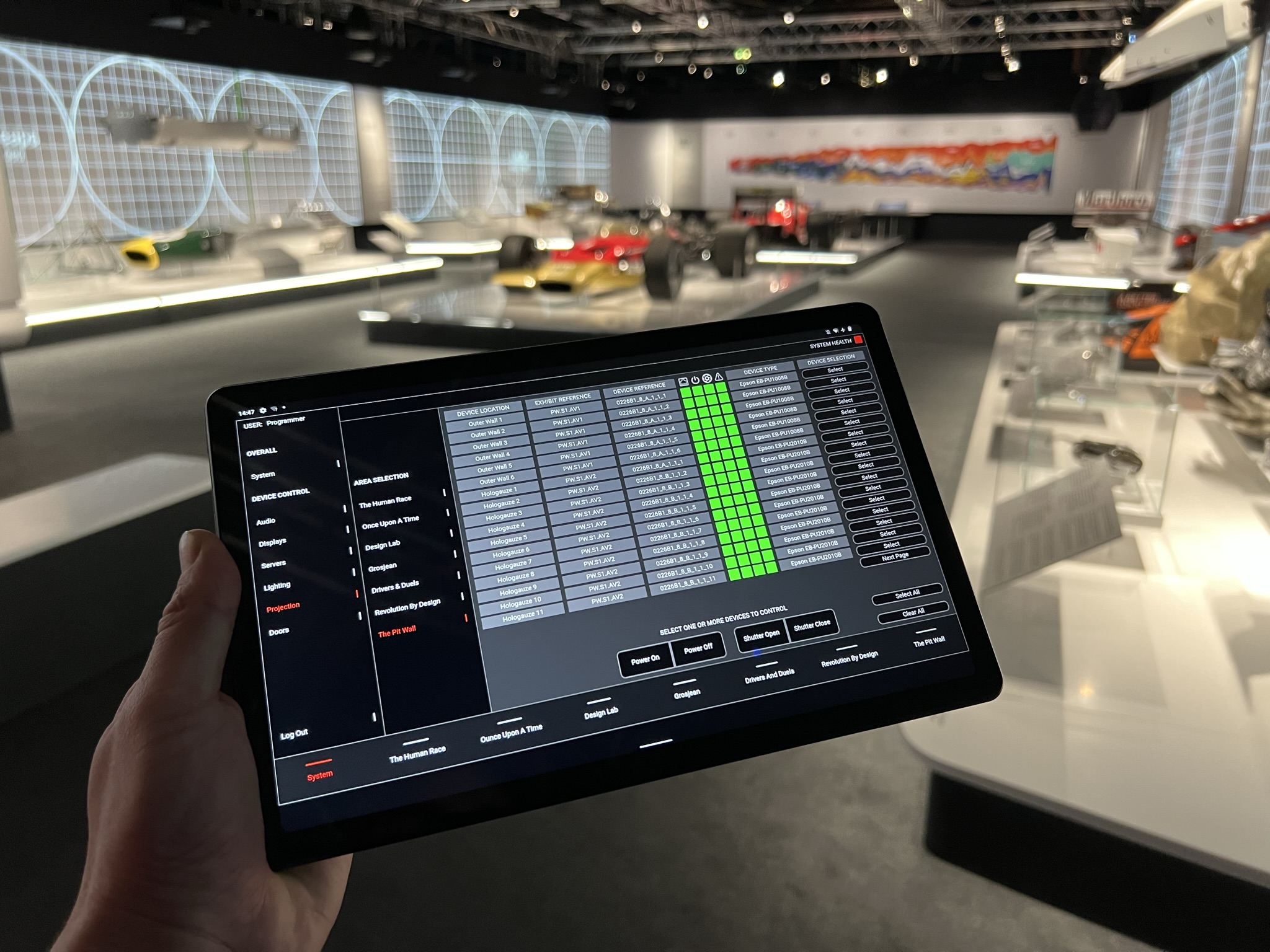 A hand holds a black tablet displaying a technical "System Control" interface in a dark gallery. The screen shows a detailed table of "Projection" devices for different exhibit areas like "The Pit Wall" and "Revolution By Design."