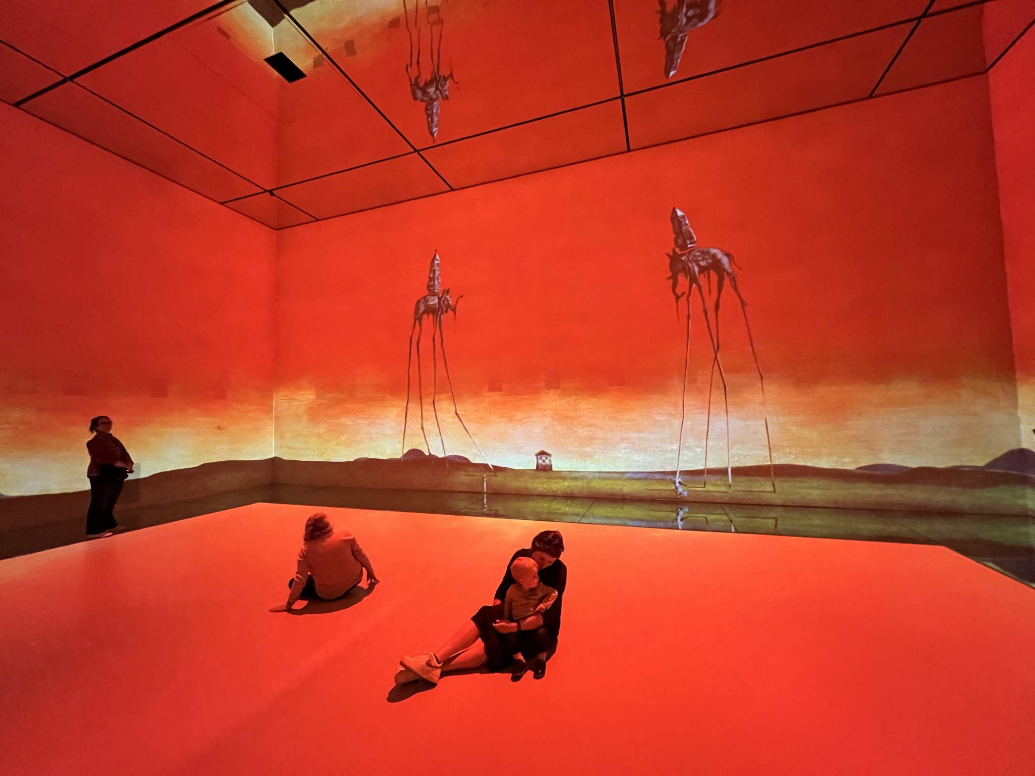 A vast, immersive art gallery bathed in deep orange and red light. A projection of Salvador Dalí’s "The Elephants" covers the walls, showing two long-legged elephants carrying obelisks against a desert horizon. Three people are sitting on the glowing floor