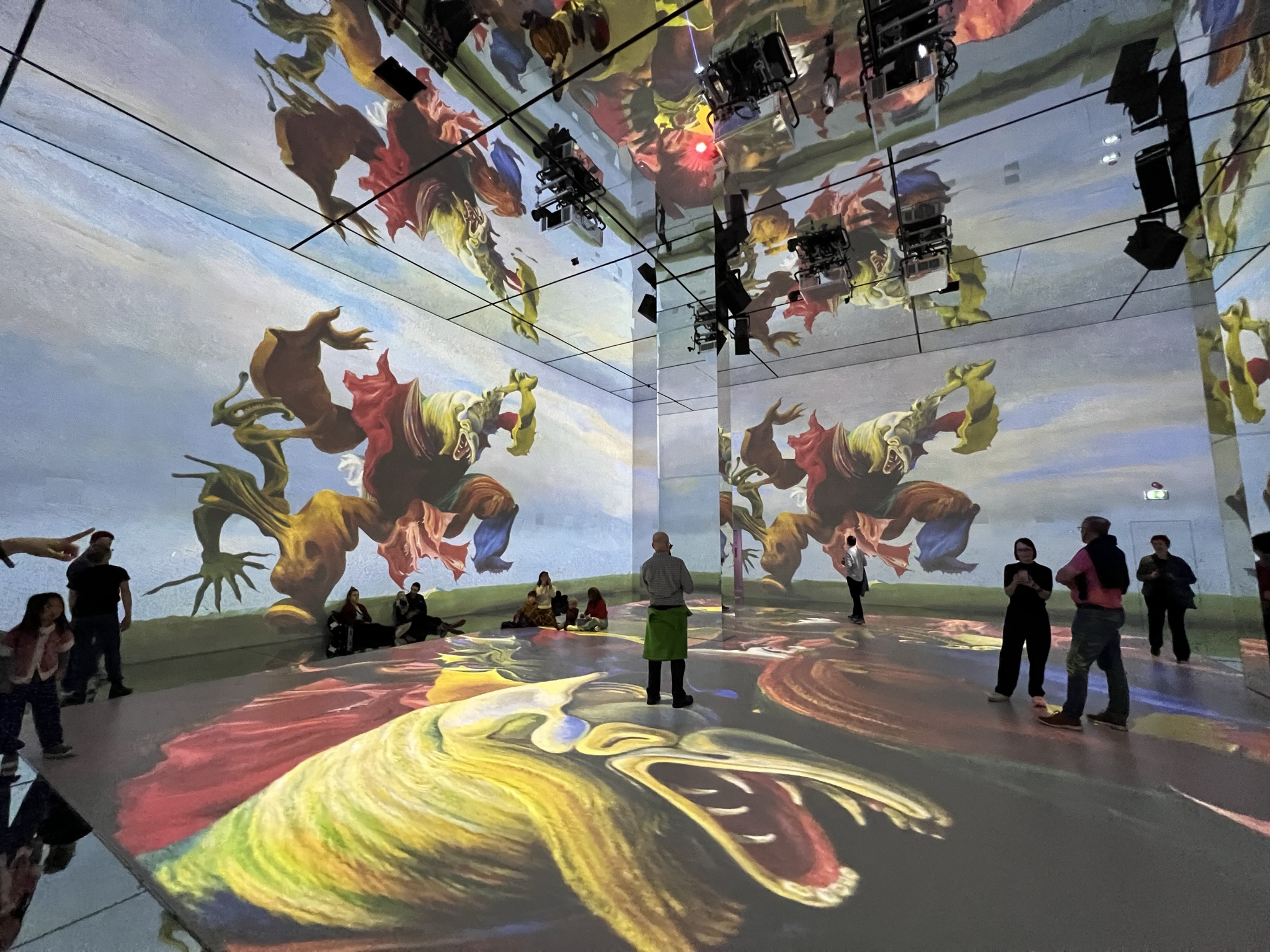 An expansive digital art installation featuring a vibrant, surrealist painting of a dancing figure projected across the walls and mirrored floor. The room is filled with several visitors who appear small against the tall, floor-to-ceiling projections.