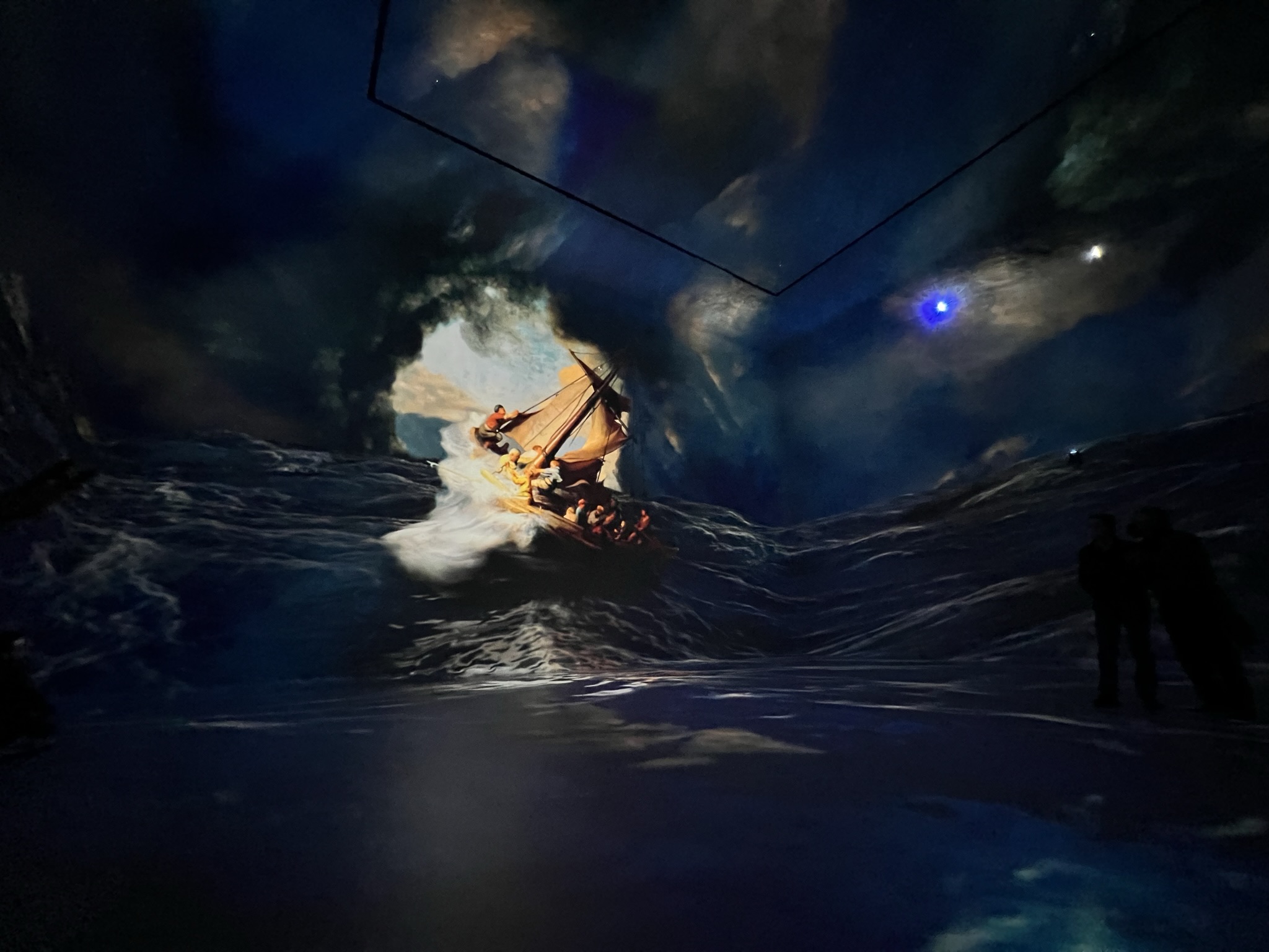 A dark, atmospheric room where a dramatic digital projection of a stormy sea covers every surface. In the center, a ship filled with people struggles against massive, crashing white waves under a swirling, dark blue and gold sky.