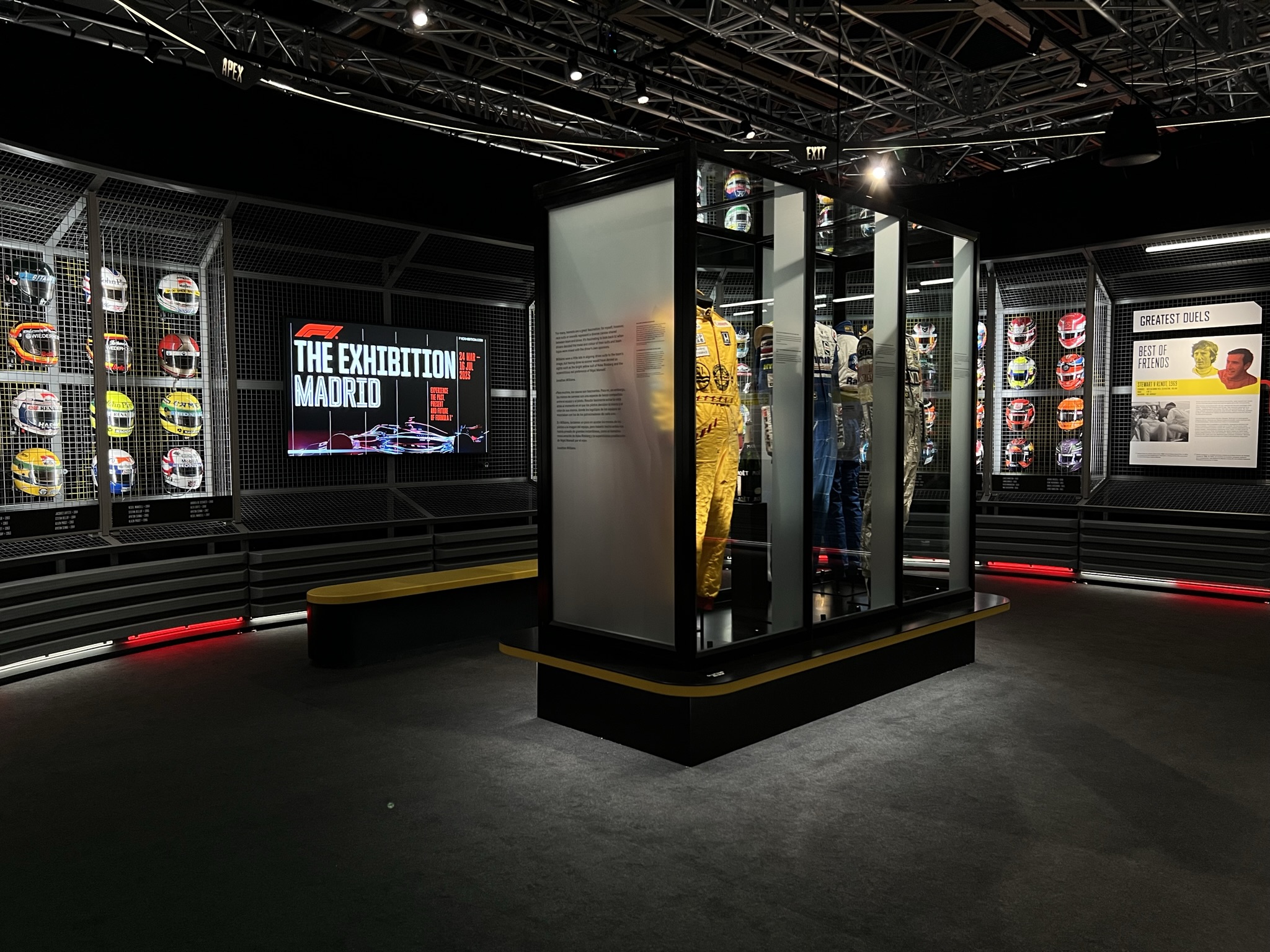 An exhibit area within "The Exhibition Madrid" featuring Formula 1 memorabilia. In the center is a tall glass display case containing a yellow racing suit. To the left, a wall-mounted grid displays a variety of colourful racing helmets.
