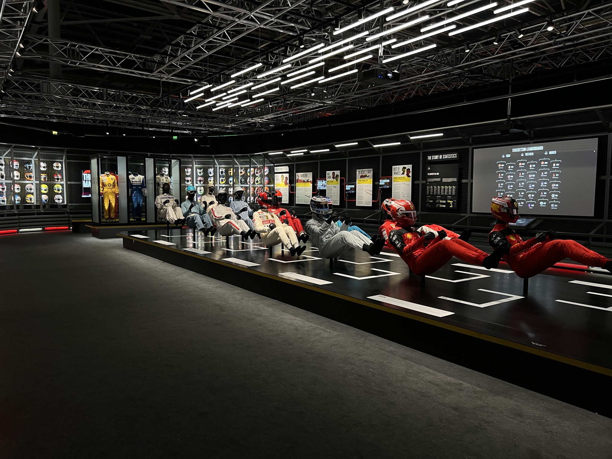 A wide shot of a museum exhibit titled "The Story of Statistics." A row of mannequins dressed in various colored racing suits and helmets are seated in a reclined, driving position on a long black platform