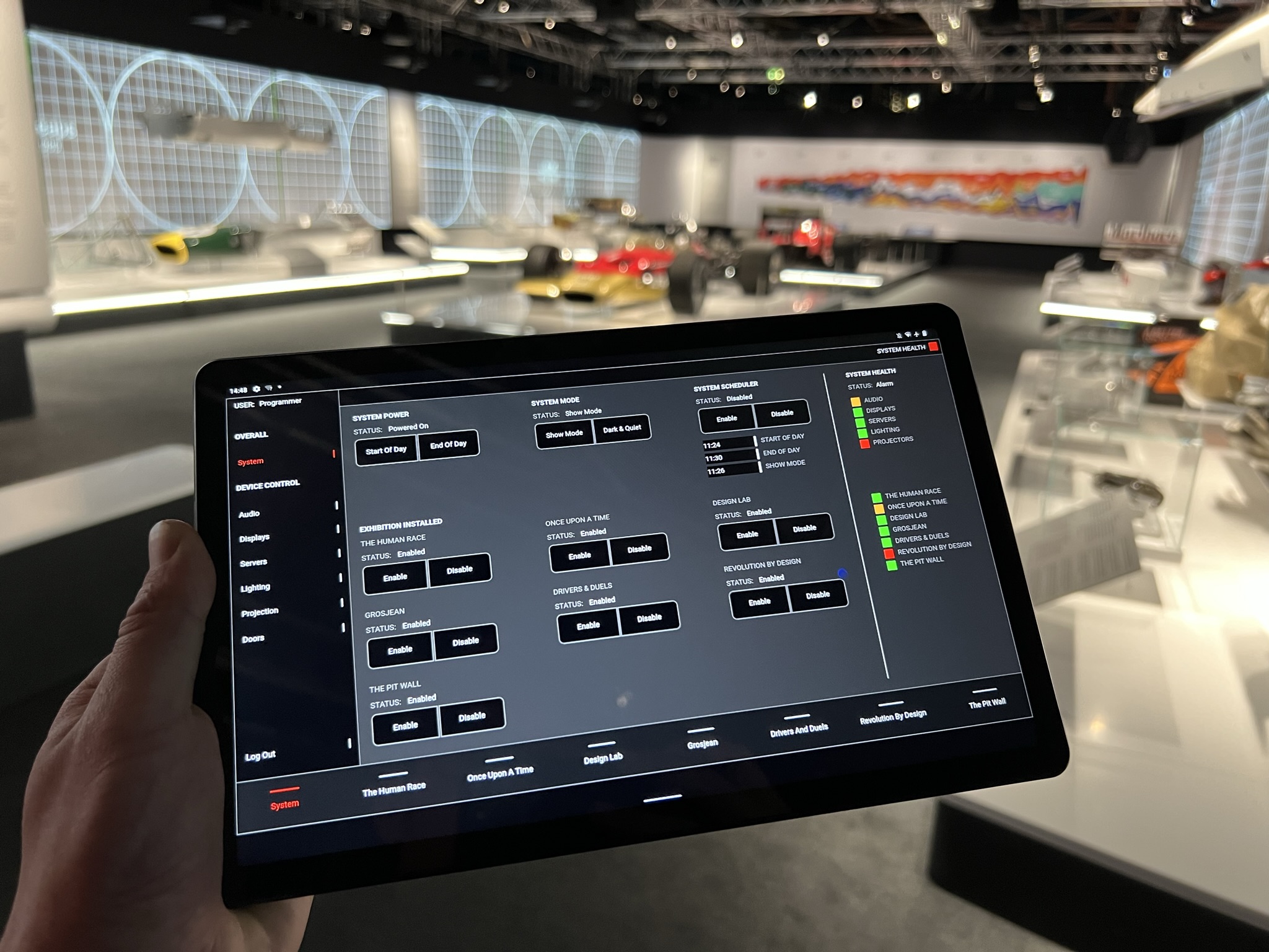 A hand holds a tablet displaying an "Overall System" dashboard for an exhibition. The interface includes toggle buttons to enable or disable different exhibit sections. A "System Health" sidebar shows green status lights for audio, displays, and servers.