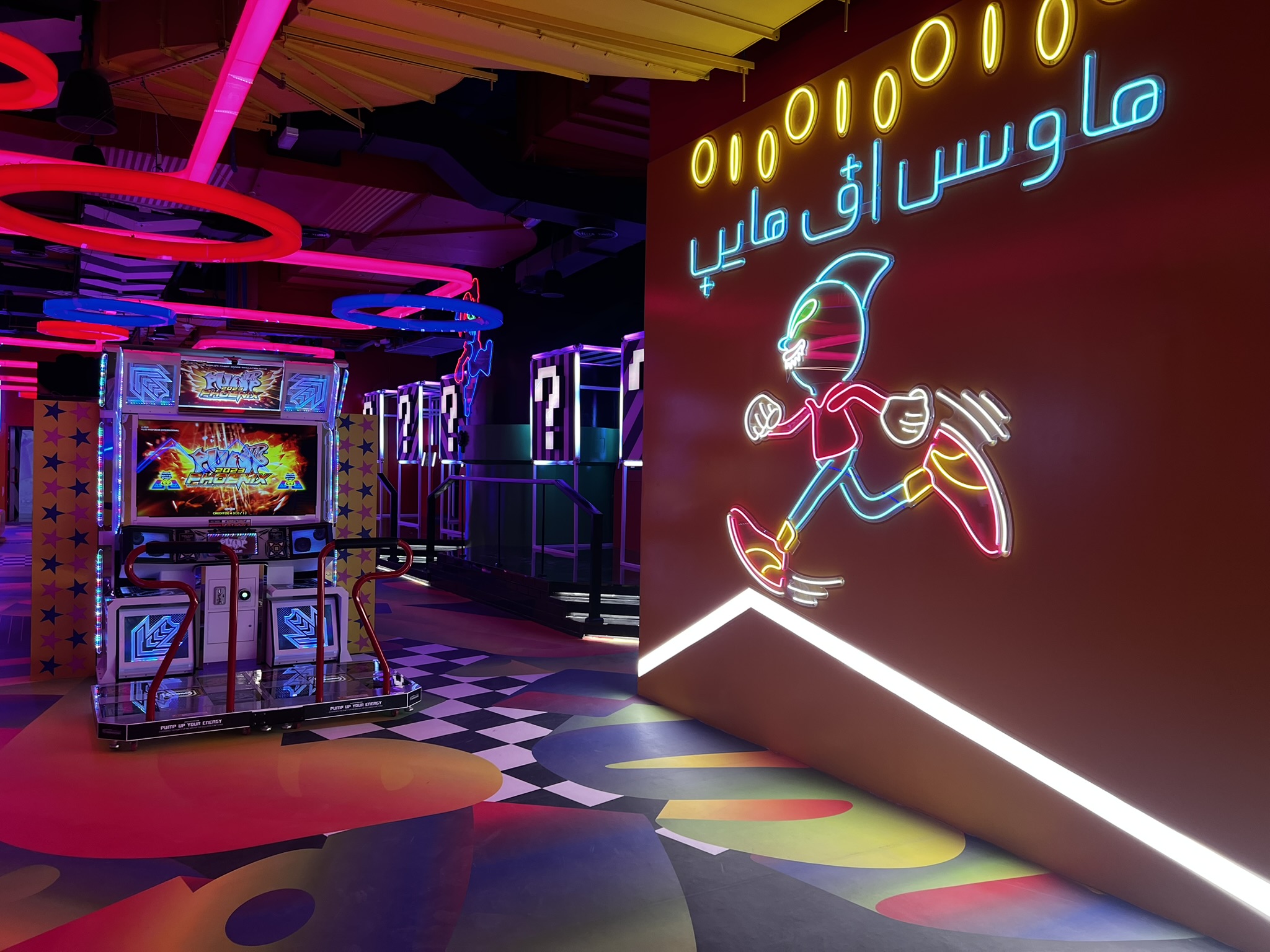 A high-energy arcade room featuring a "Pump It Up Phoenix" dance machine on a colorful patterned floor. To the right, a large orange wall features a neon light display of a running cartoon character
