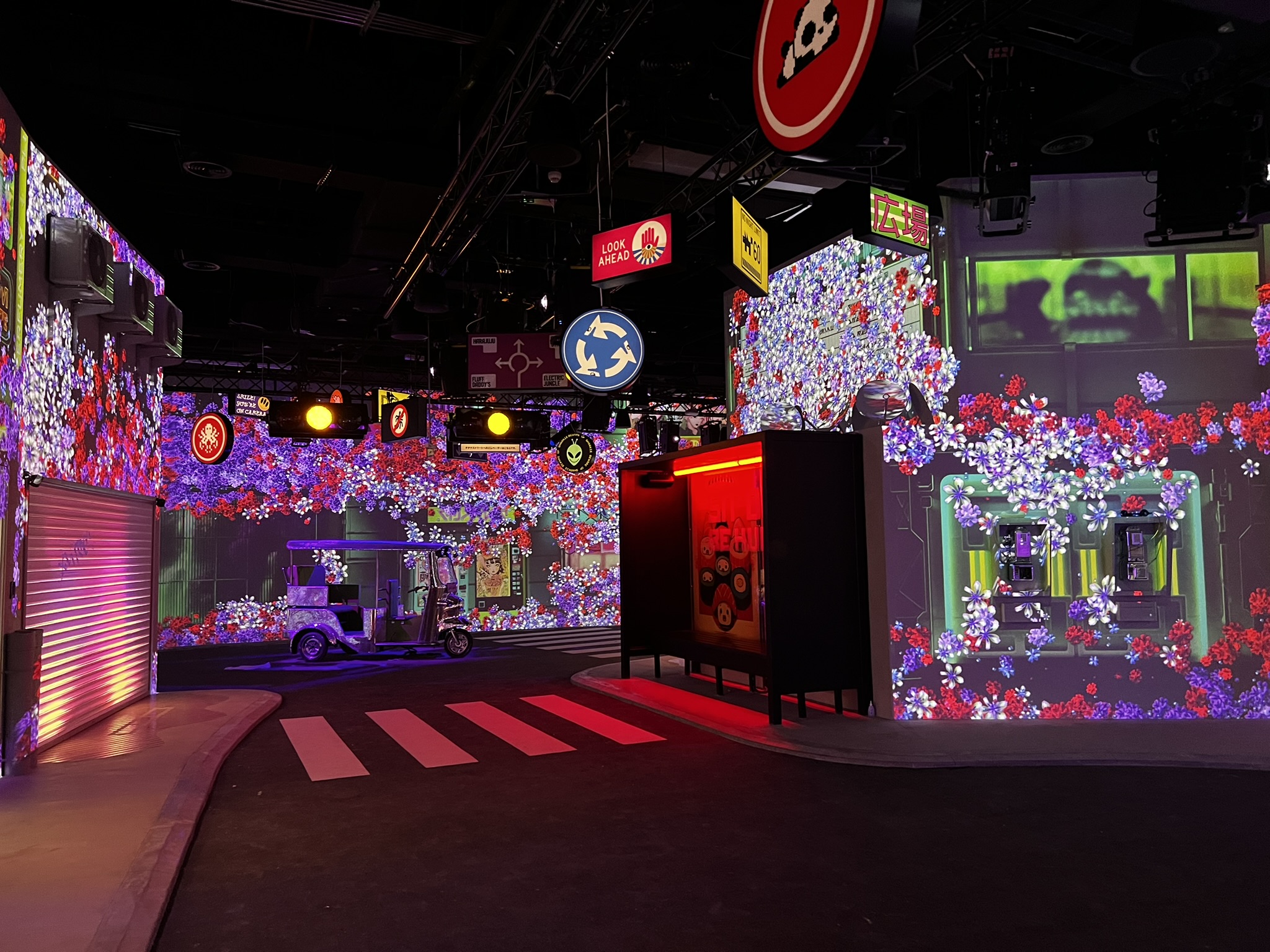 A life-sized indoor street scene immersive art installation. The walls are covered in digital projections of dense purple and red flowers.