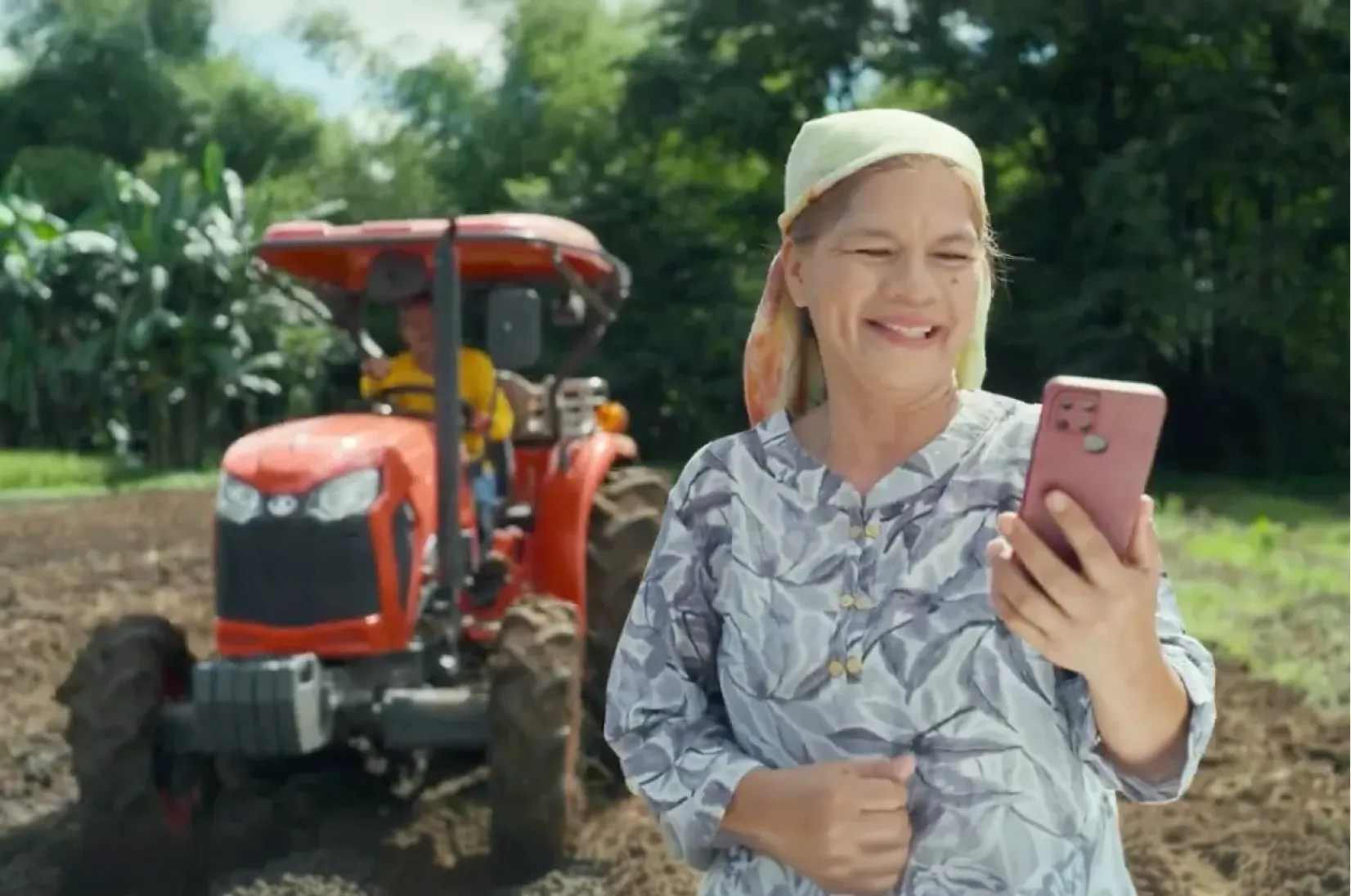 Smiling woman in patterned shirt and headscarf using a pink smartphone in a field with an orange tractor and farmer in the background.