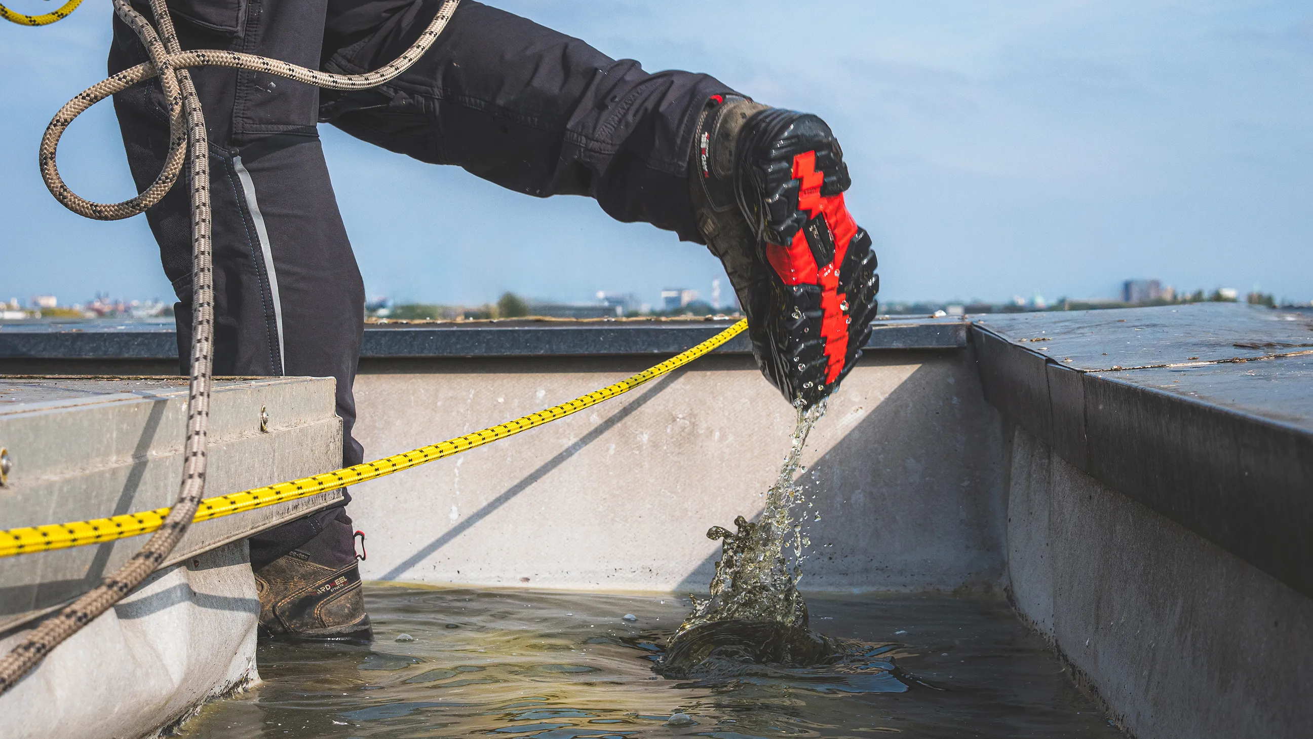 Photo prise au niveau des jambes, ou une chaussure marche dans l'eau.
Il y a de l'eau sur un toit et un cordiste vient régler le soucis.