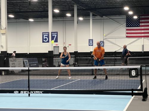 Two people playing pickleball indoors on court 6, with an American flag and courts 5,7,8,9 visible in the background.