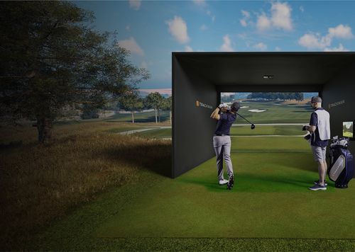 A golfer swinging a club inside a golf simulator setup with a caddy standing nearby holding a golf bag.