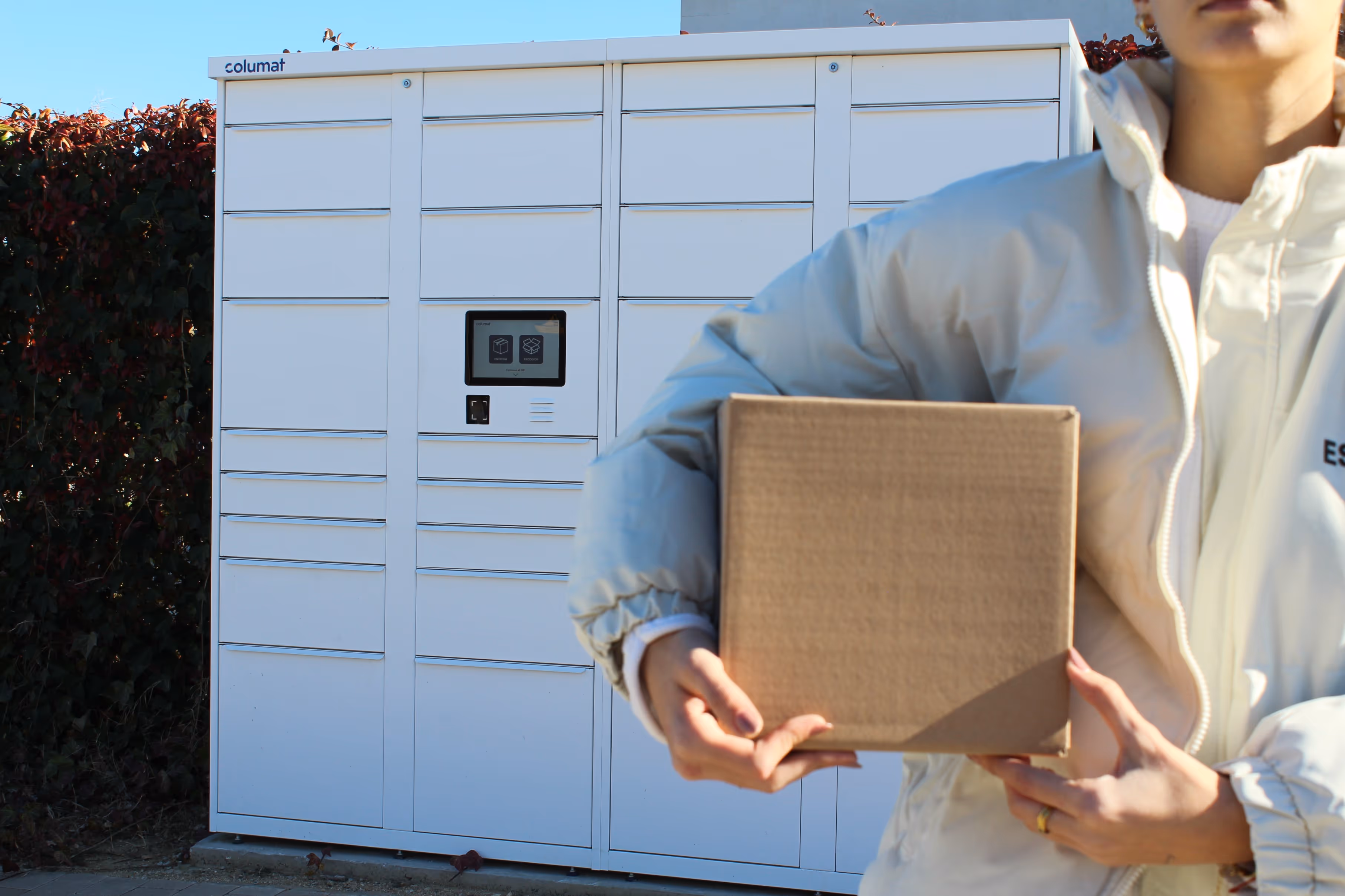 Person in a light-colored jacket holding a cardboard box in front of a white Columat parcel locker outdoors.