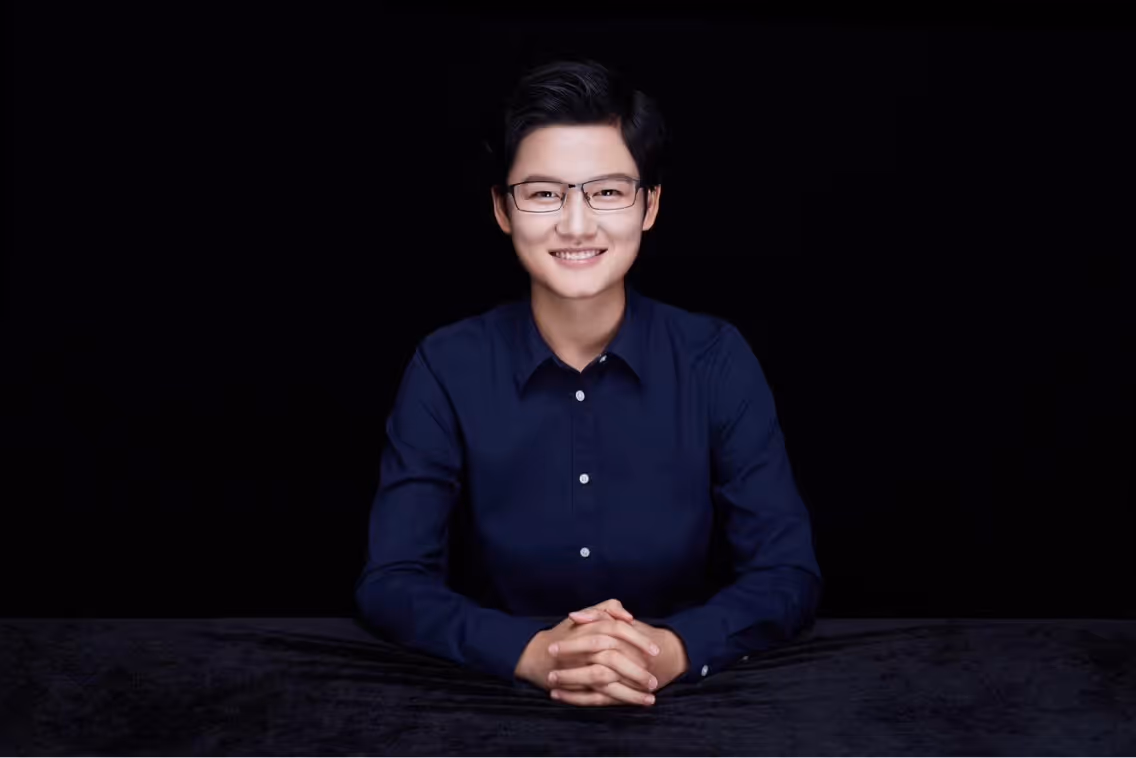 Smiling young person with short dark hair and glasses, wearing a dark blue button-up shirt, sitting with hands clasped on a black surface against a black background.