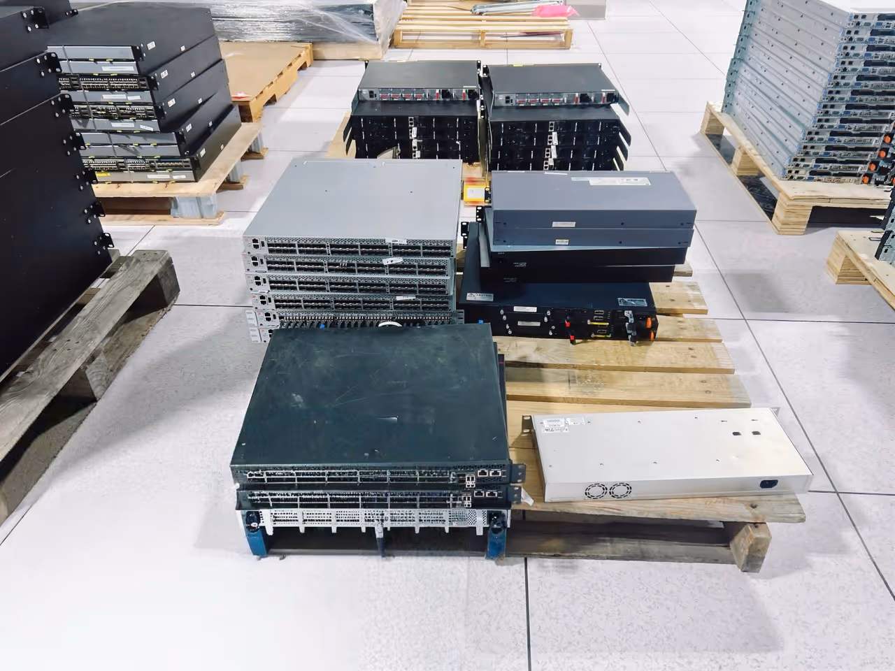 Stacks of network switches and servers on wooden pallets in a warehouse with tiled flooring.