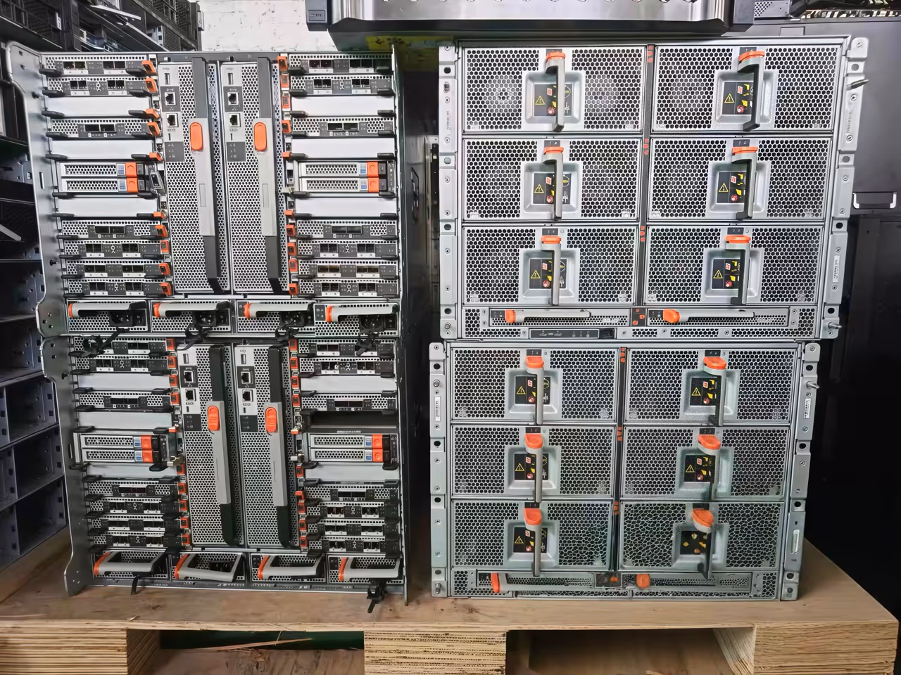 Two server racks with multiple units featuring metal mesh fronts and orange handles, placed on a wooden pallet inside a server room.