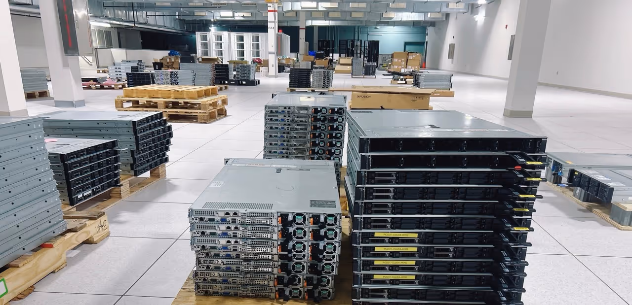 Stacks of servers and computer hardware arranged on wooden pallets in a large, empty warehouse with tiled flooring and overhead lighting.