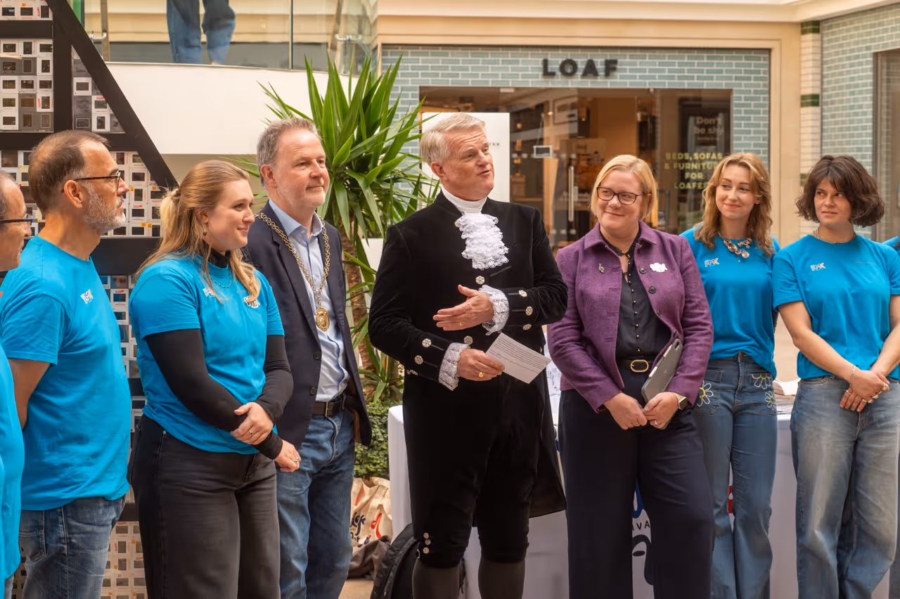 Group of people standing indoors, including a man dressed in formal ceremonial attire speaking, surrounded by casually dressed individuals and a man wearing a mayoral chain.