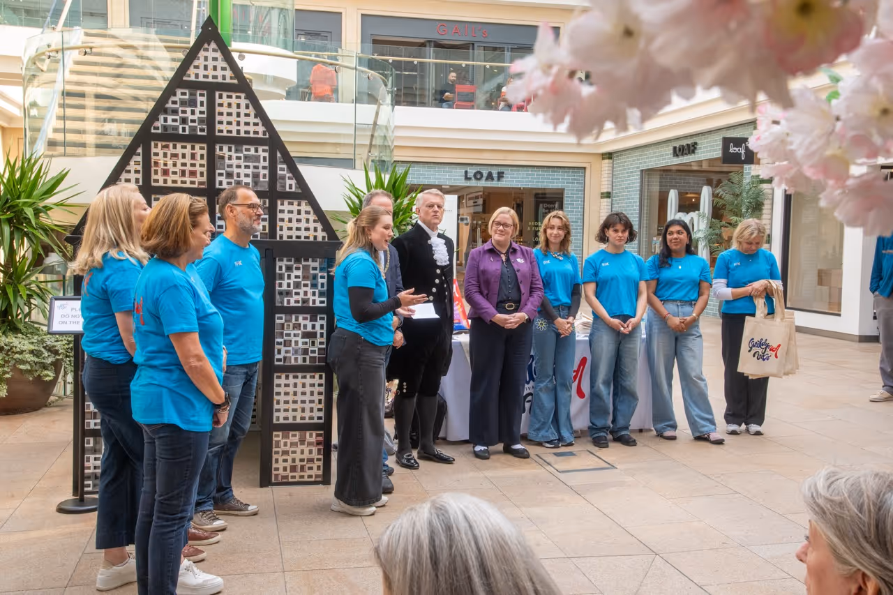 Group of people, mostly wearing blue shirts, standing in a semi-circle indoors near a black triangular display and table with a white cloth in a shopping area.