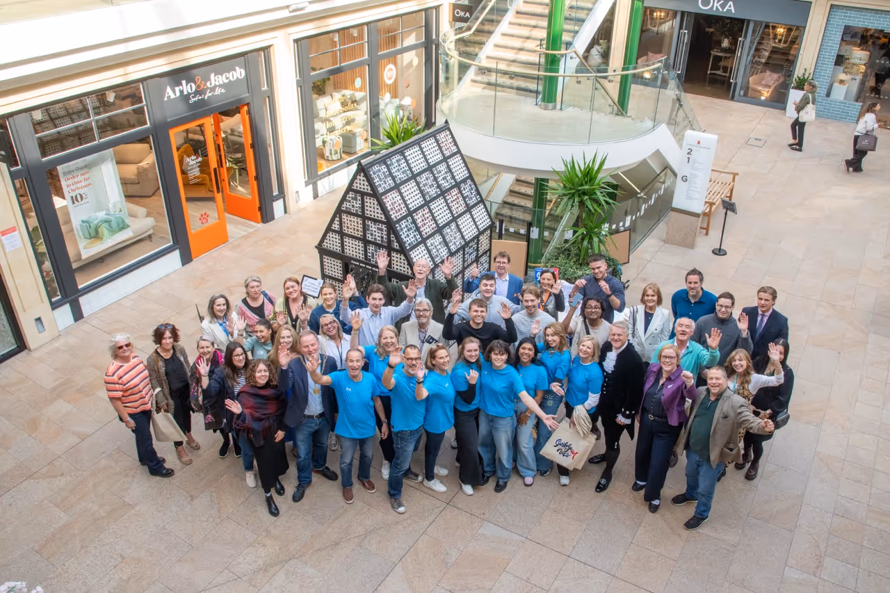 Group of Guildford Festival of the Arts team members smiling and waving in a shopping mall near a display shaped like a house.