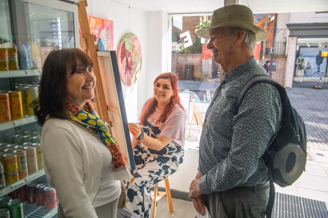 Young woman painting on a canvas inside a bright studio, smiling and engaging with an older man wearing a hat and a woman with a colorful scarf.