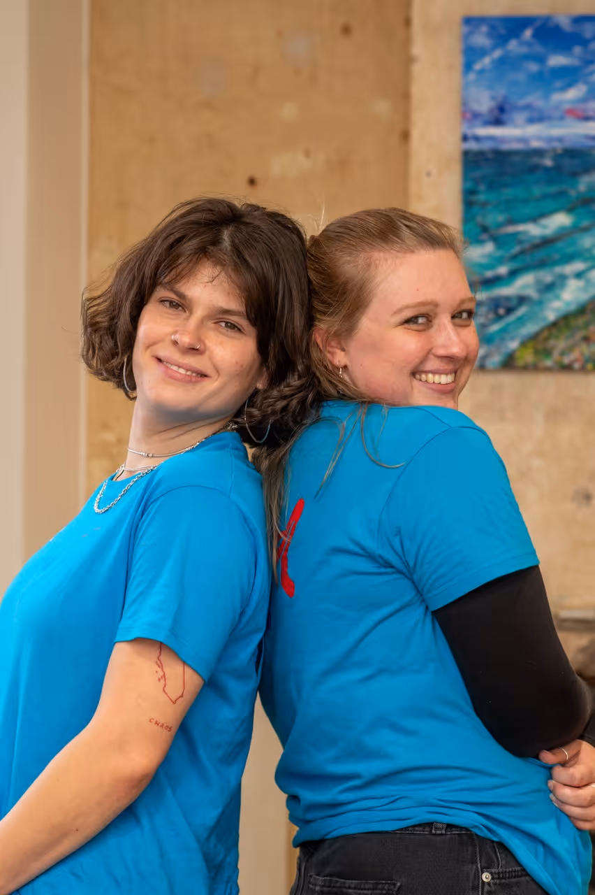 Two young women in blue t-shirts smiling and leaning back against each other indoors with a colourful painting on the wall.