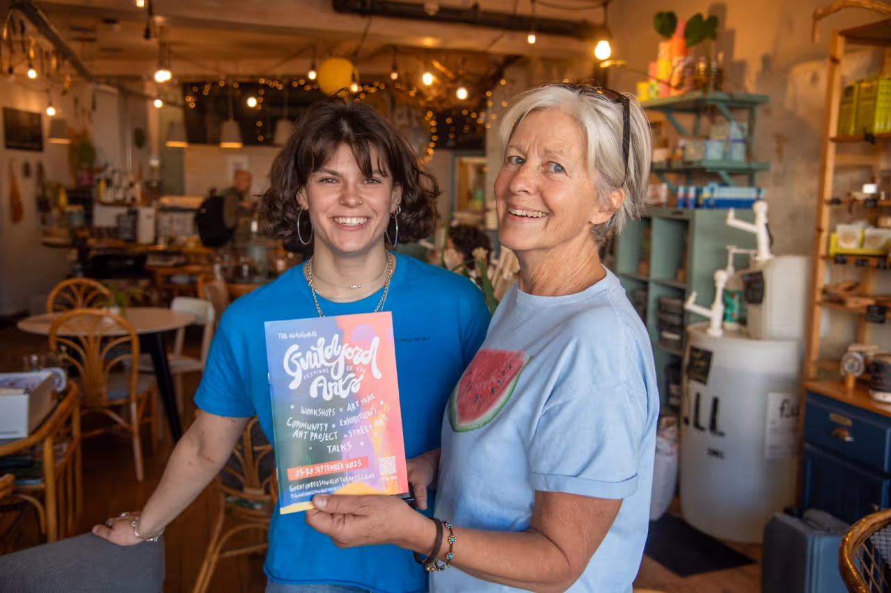 Two smiling women in a cozy café holding a colorful Guildford Festival of the Arts 2025 flyer.