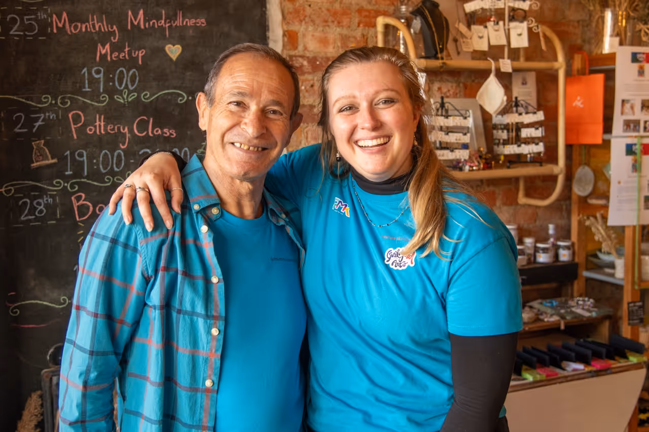 Smiling man and woman wearing blue shirts, posing together in a shop with a blackboard and display shelves in the background.