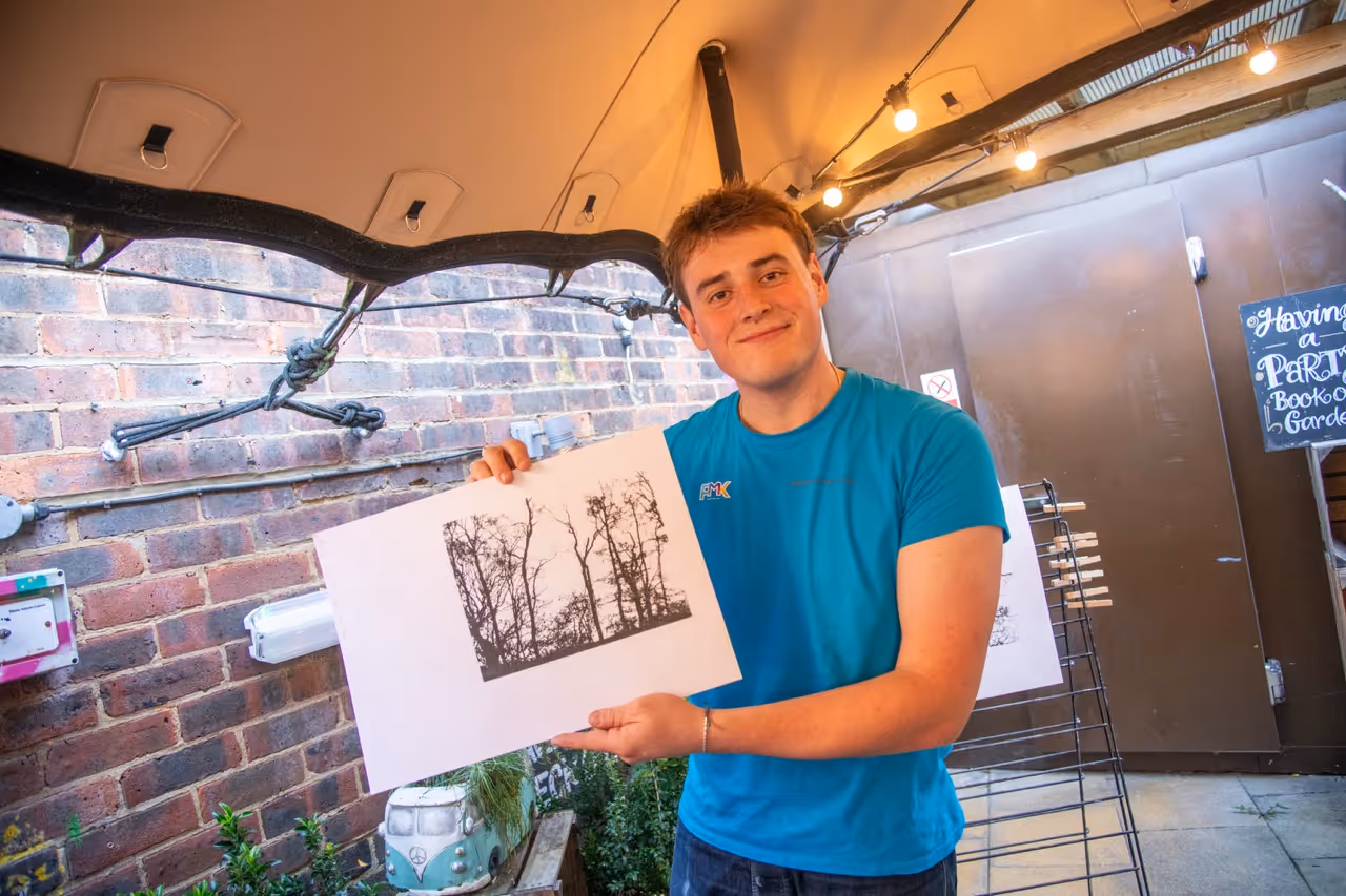 Young man wearing a blue Guildford Festival of the Arts t-shirt smiling and holding a screen printed artwork.
