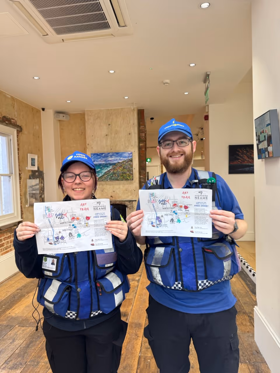Two smiling town rangers wearing blue uniforms and caps holding up maps of the Art Trail inside a brightly lit room with wooden floors and artwork on the walls.
