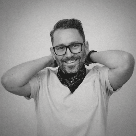 Smiling man with glasses, a beard, and a bandana around his neck posing with hands behind his head.