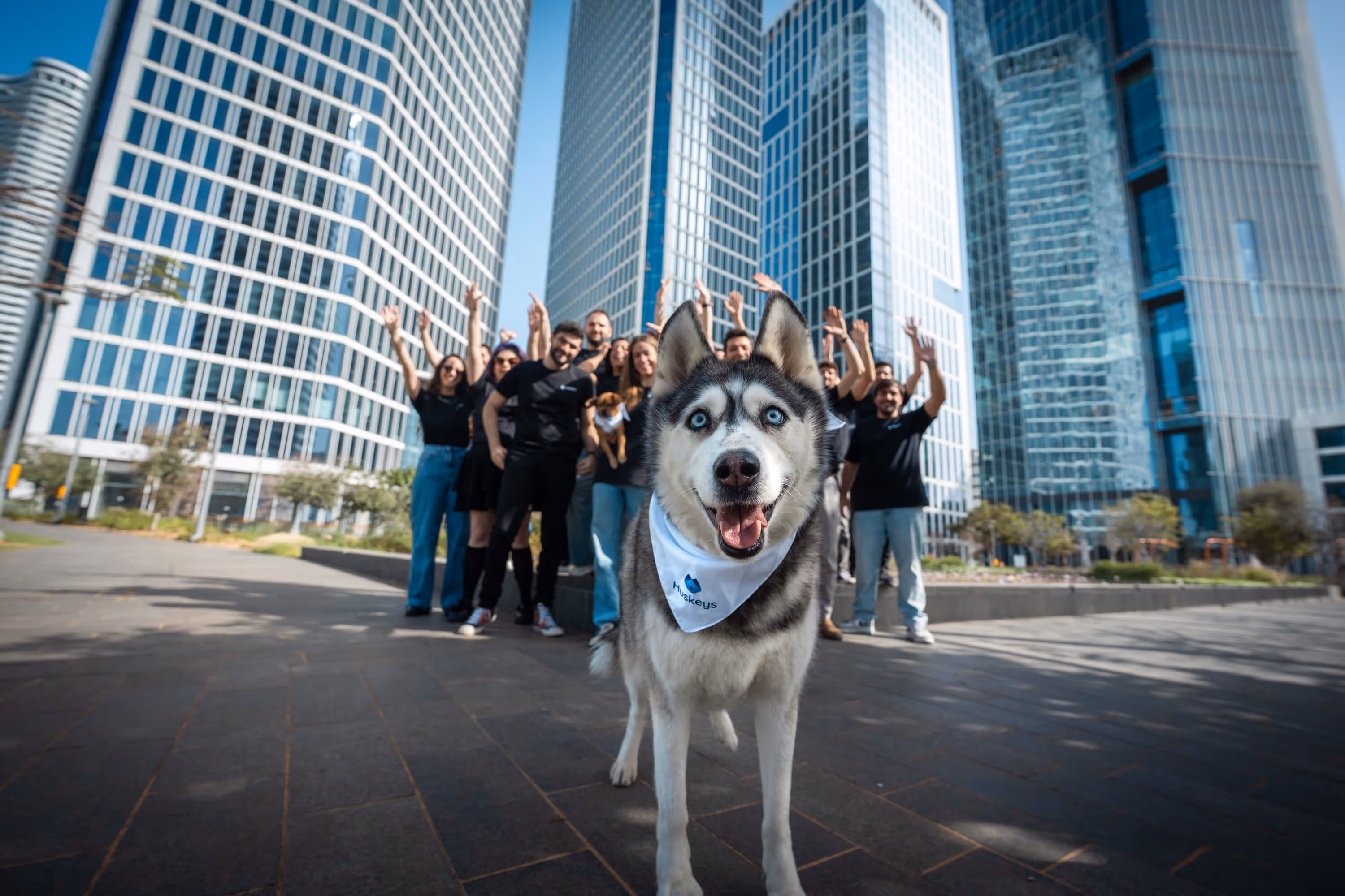 Happy husky dog wearing a white bandana stands in the foreground with a group of smiling people and tall modern office buildings behind.