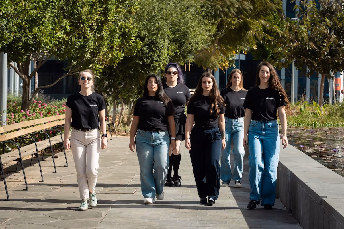 Group of six women wearing black Husqvarna t-shirts walking confidently outdoors along a paved path with trees and water lilies in the background.