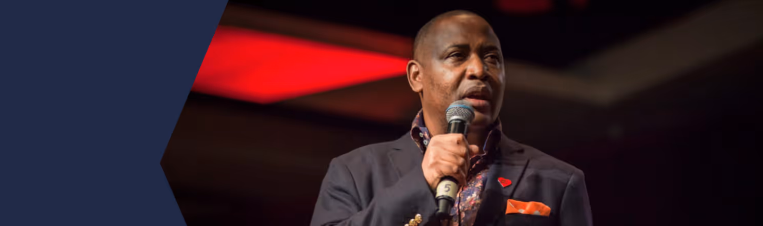 Man speaking into a microphone against a dark background with red lighting.