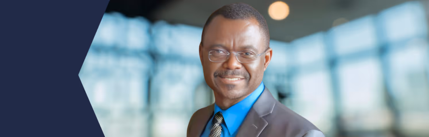Smiling man wearing glasses, a blue shirt, patterned tie, and dark suit jacket against a blurred indoor background.