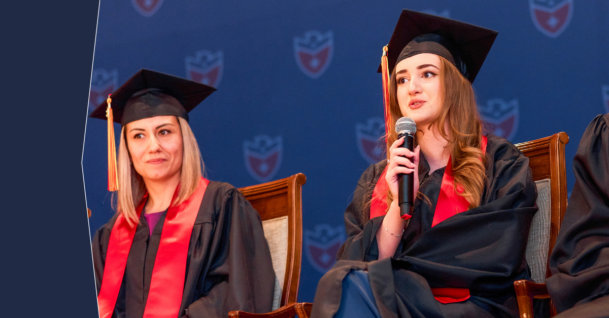 Two female graduates in caps and gowns sitting on stage, one speaking into a microphone.