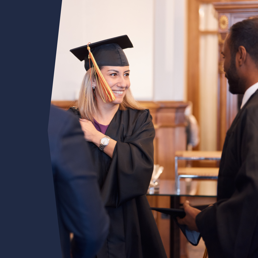 Smiling graduate woman wearing a black cap and gown during a graduation ceremony inside a formal venue.