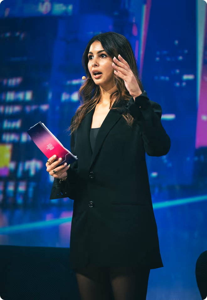 Portrait d'une femme brune avec un blazer noir posant devant un fond sombre, main levée vers le visage.