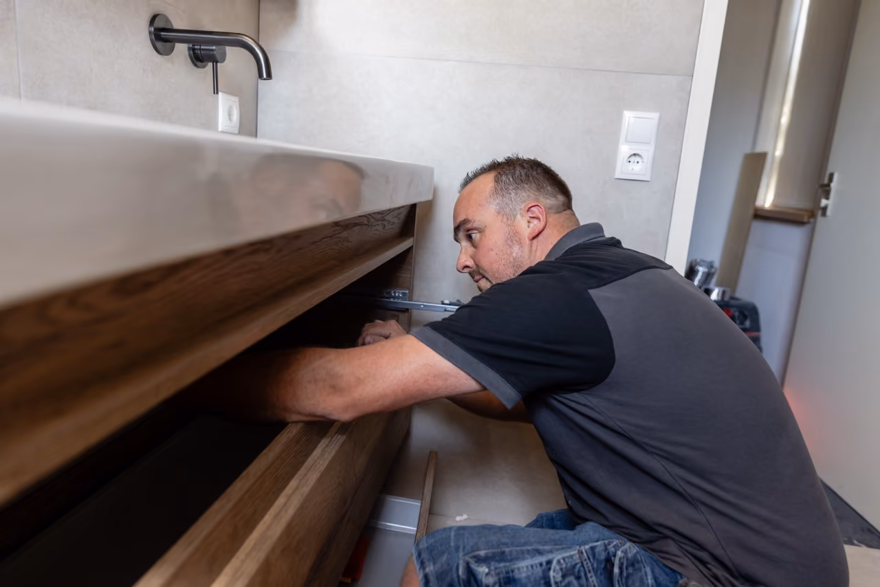 Installateur van Marboma monteert een houten badmeubel onder een wastafel met zwarte kraan