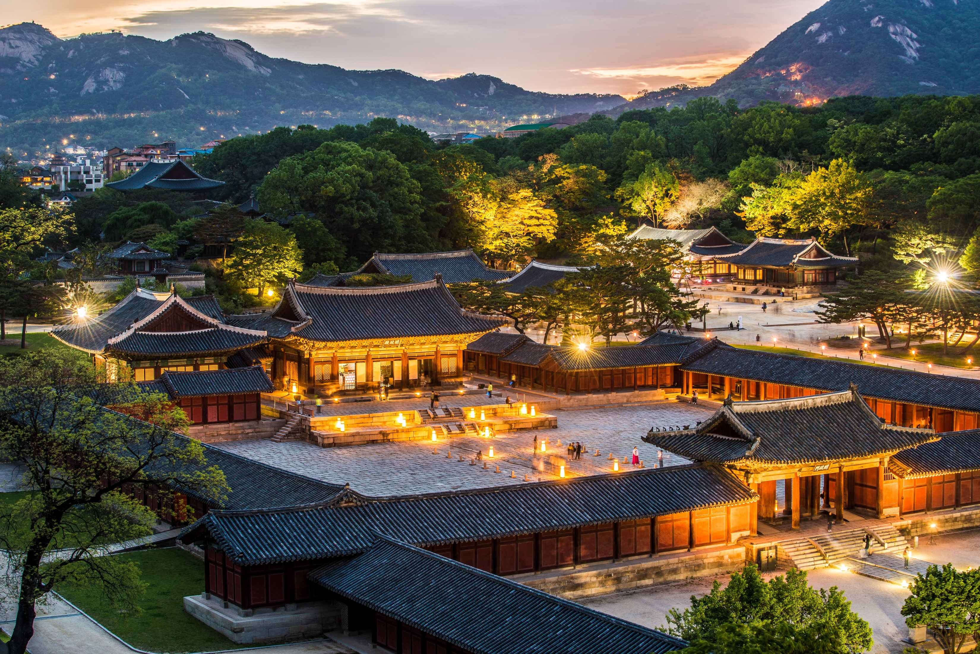 Illuminated traditional Korean palace buildings at dusk with mountains and trees in the background.