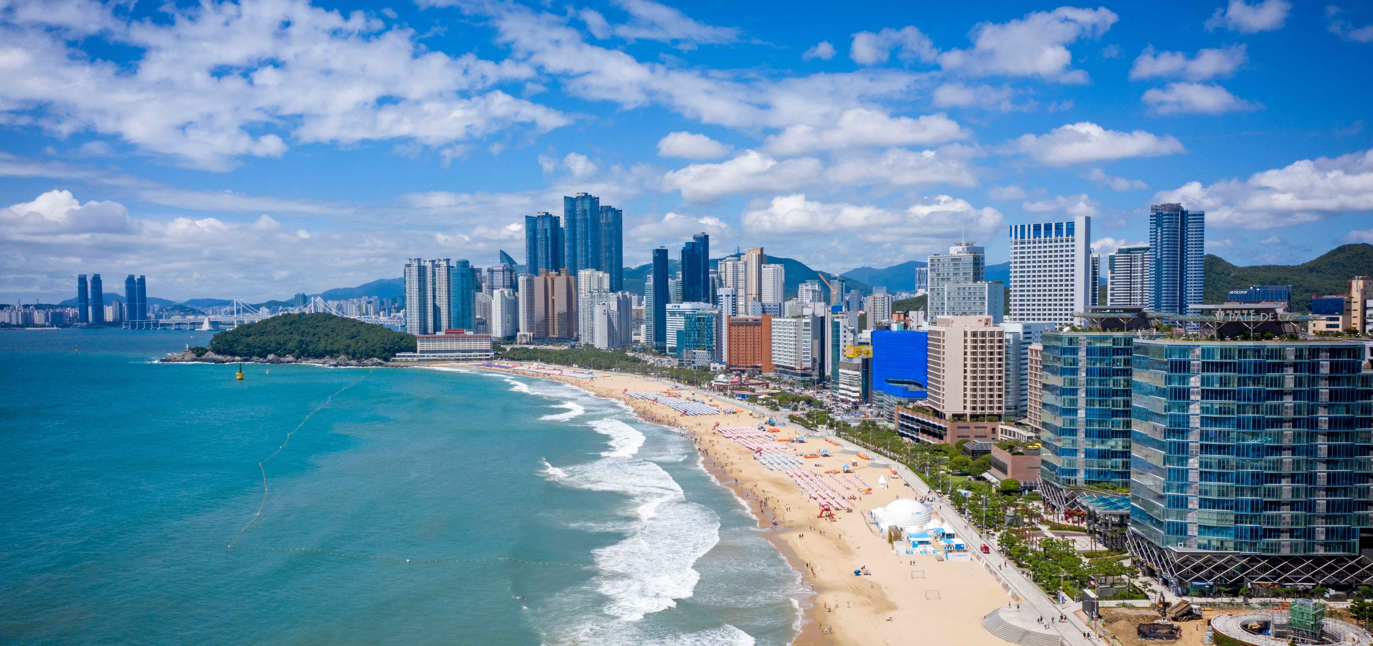 Panoramic view of a sandy beach with umbrellas and people, turquoise ocean waves, and a city skyline with high-rise buildings under a partly cloudy blue sky.