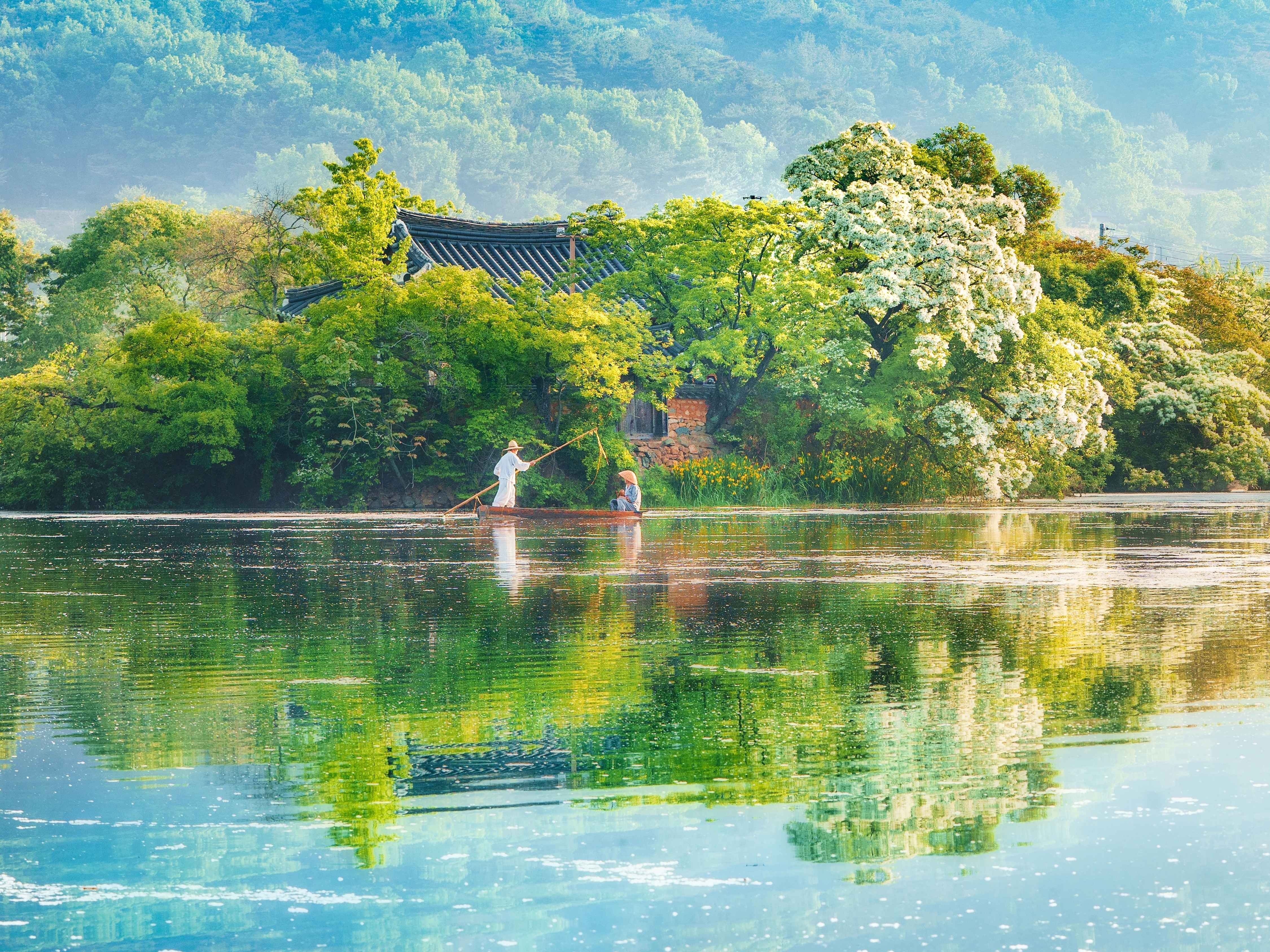 Two people on a small boat fishing on a calm lake with lush green trees and a traditional building in the background.