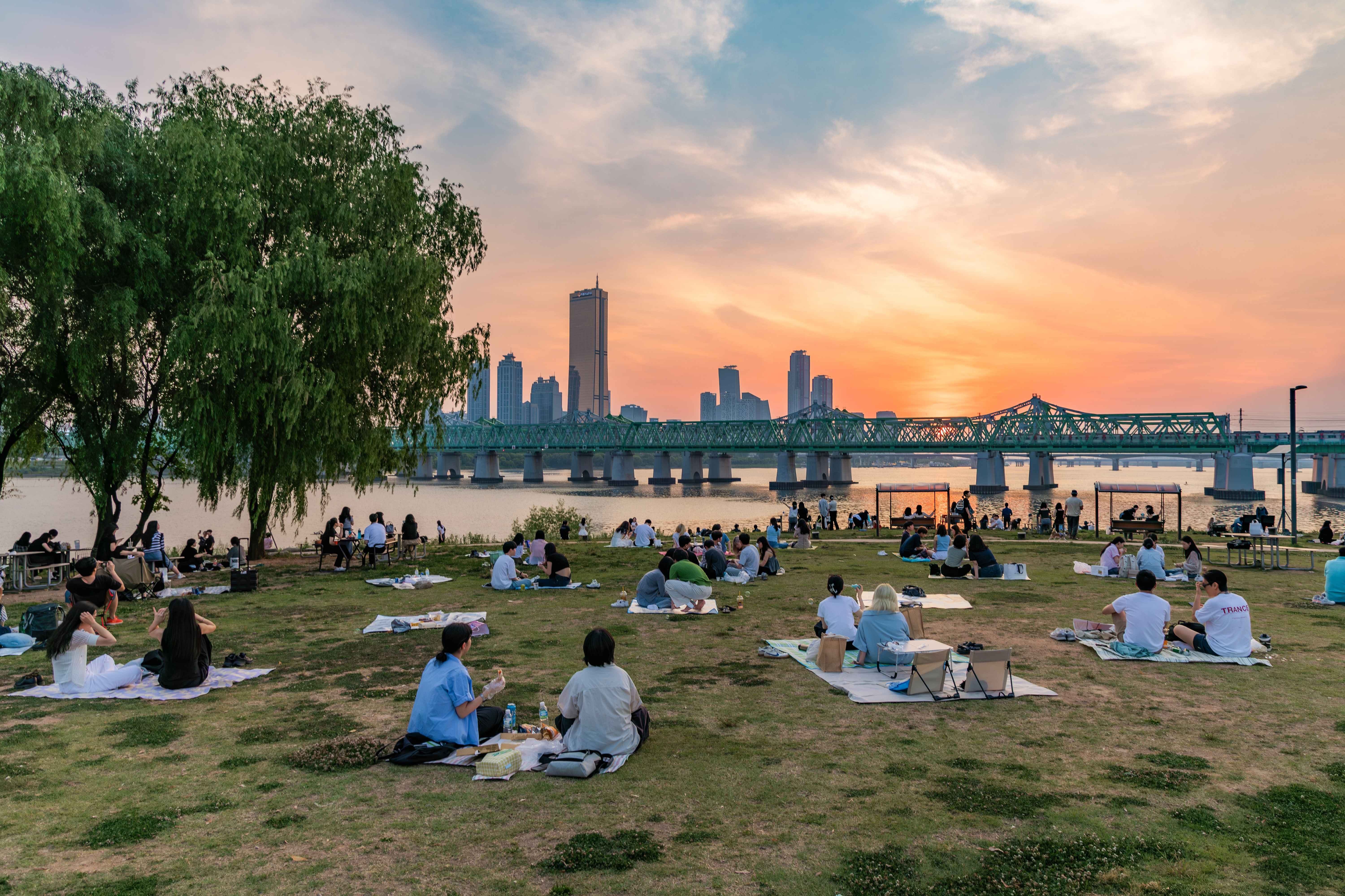 People having picnics on a grassy riverside park at sunset with a green steel bridge and city skyline in the background.