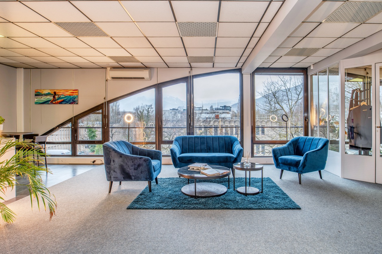 Salon moderne avec canapé bleu, deux fauteuils assortis, tables basses rondes et grandes fenêtres offrant une vue sur des arbres et des montagnes.