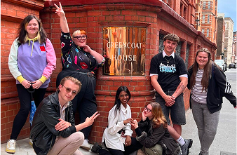Group of six young people posing and smiling in front of a brick wall with a golden plaque reading 'FRENGCOAT HOUSE.'